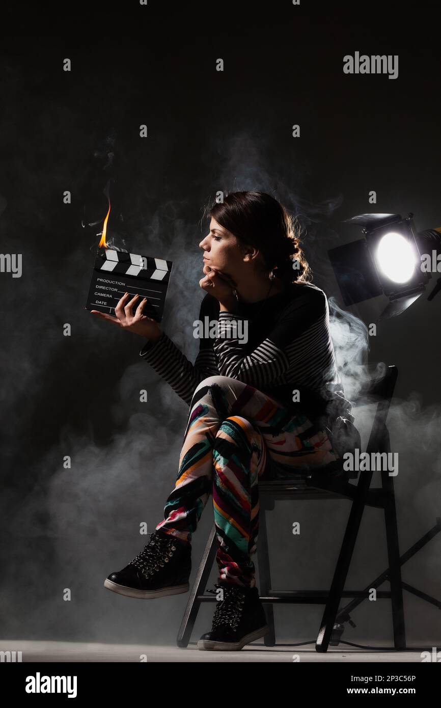 Female movie director on set with smoke background. Girl holding clapperboard on fire. Stock Photo