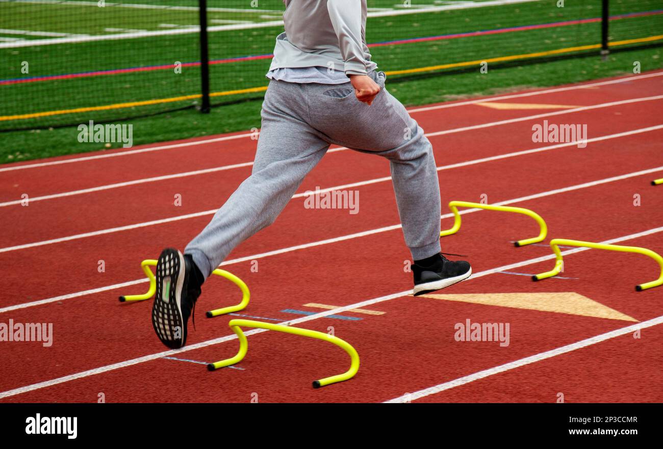 Side view of a high school boy running the wicket drill over yellow mini banana hurdles on a red track during track practice. Stock Photo