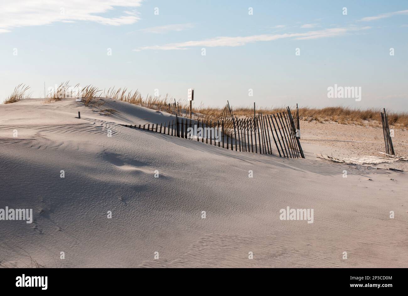 Side view of a sand dune with the sand smooth fron the wind and burying parts of the picket fence with a keep off the dubes sign in the background. Stock Photo