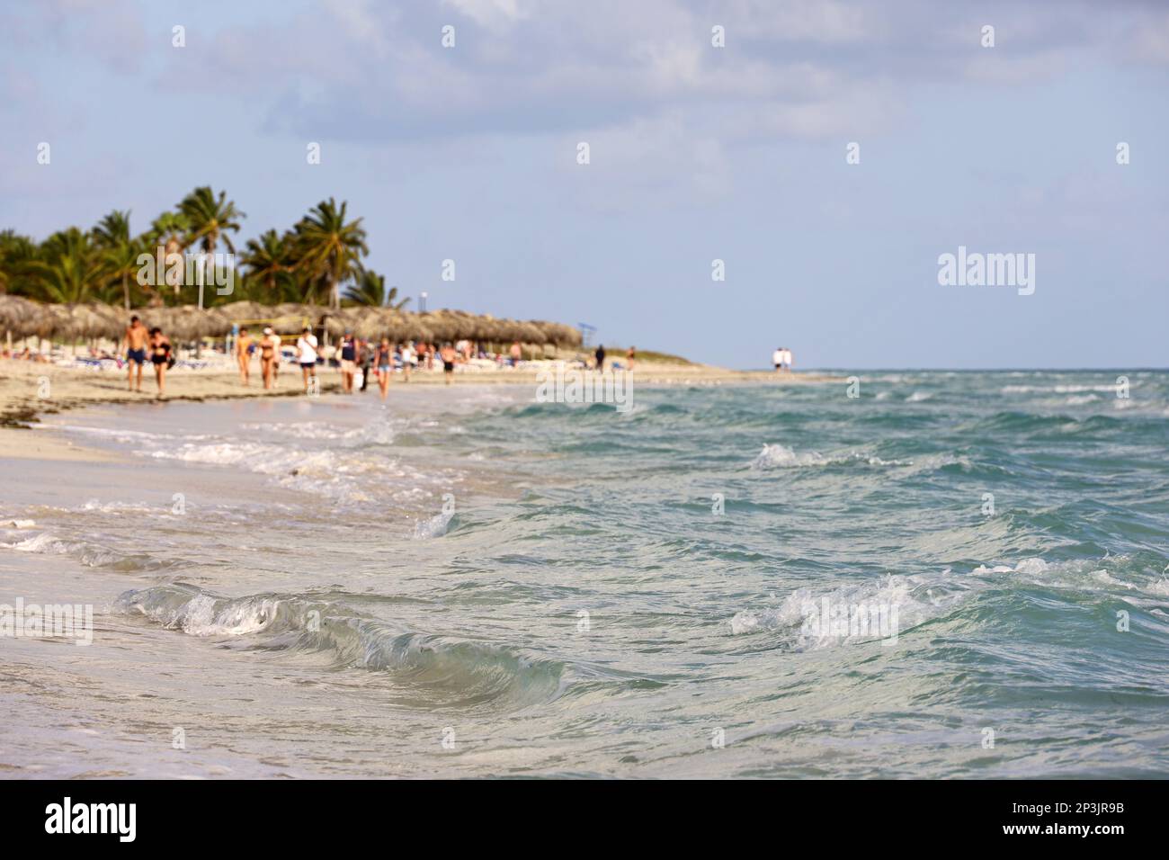 Defocused view from ocean waves to tropical beach with people, sandy coast and coconut palm trees. Caribbean resort on with transparent water Stock Photo