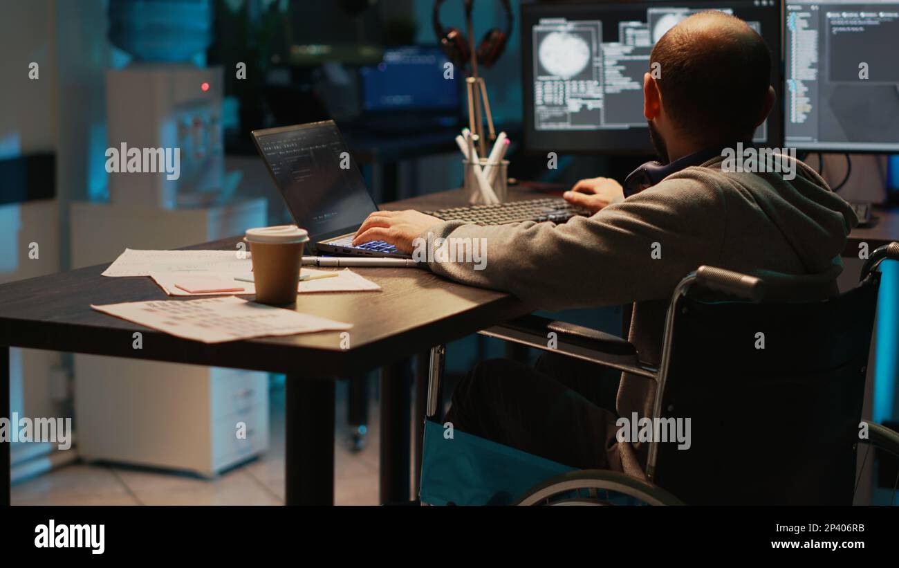Software developer with impairment working on app programming, using laptop and multiple screens. Male IT coder with chronic disability in wheelchair developing new user interface. Stock Photo