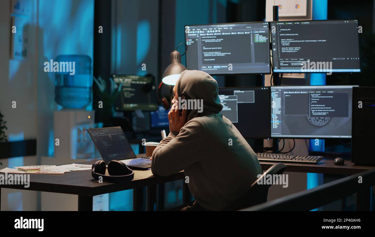 Young programmer answering phone call in office, talking on smartphone and coding user interface. Having remote conversation on mobile phone line, using html script on terminal window. Stock Photo