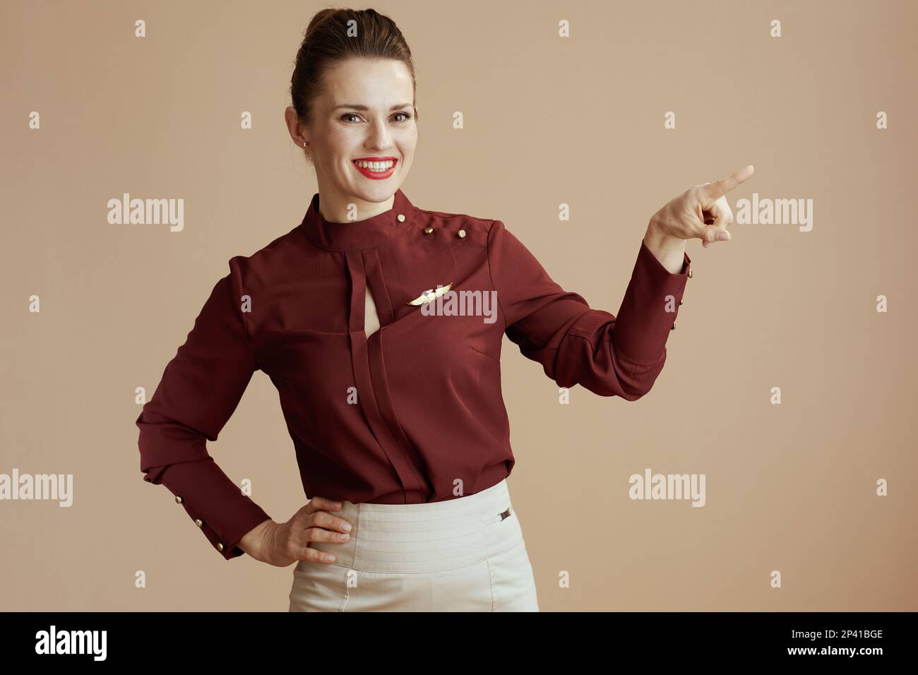 happy modern female flight attendant against beige background pointing at copy space. Stock Photo