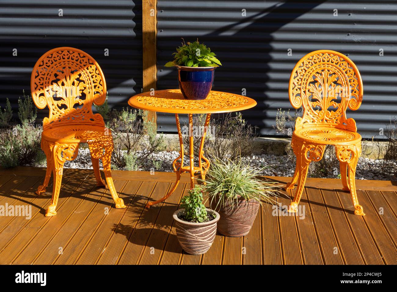 Pretty, picturesque, authentic garden chairs and table. Tidy garden pot plants. Residential property decking seating area. Bright orange cast iron set Stock Photo