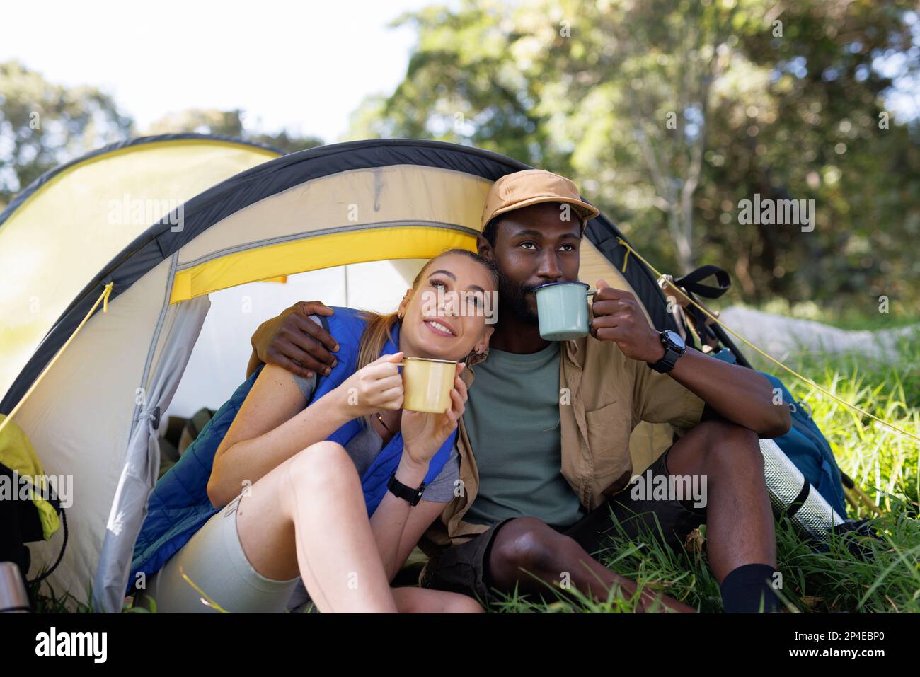 Happy, diverse couple in forest sitting in front of tent with coffee ...