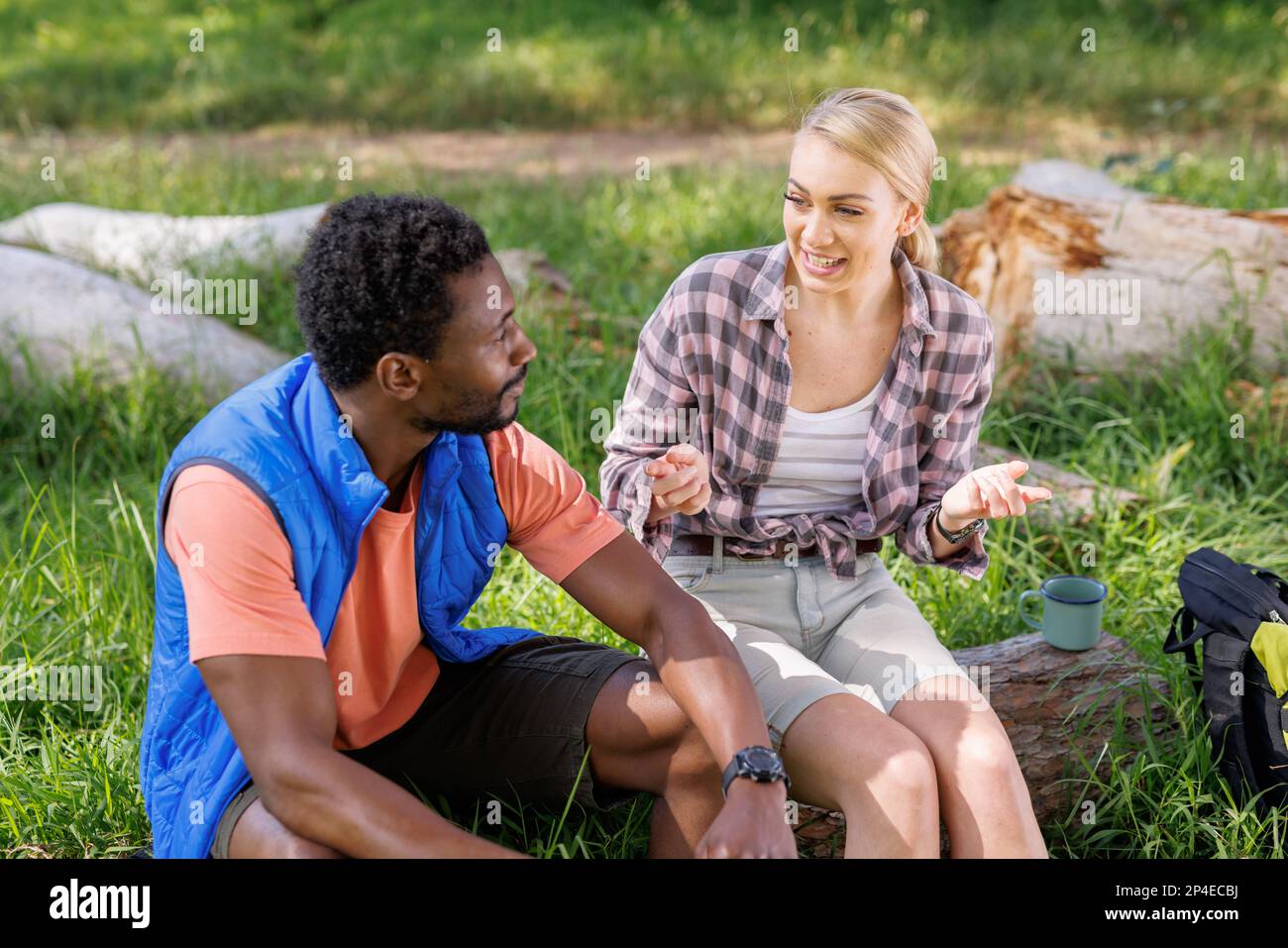 Happy, diverse couple sitting on log in sunny forest, talking Stock Photo