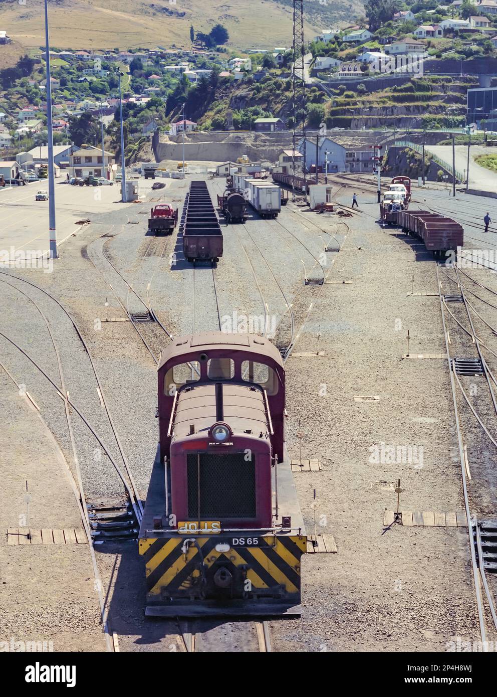 A 1981 historic image of goods trains, rail lines, and the green hills behind the goods yard in Lyttelton Harbour near the city of Christchurch on the south island of New Zealand. In the foreground is a DS class shunting locomotive DS65 that came into service in 1955 and was scrapped almost exactly a year after this shot was taken in April 1982 Stock Photo