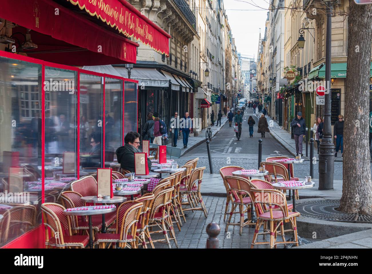 A man sits outside La Brasserie de l'Isle Saint-Louis Rue Saint-Louis en Ille stretches away in the background, Paris. Stock Photo
