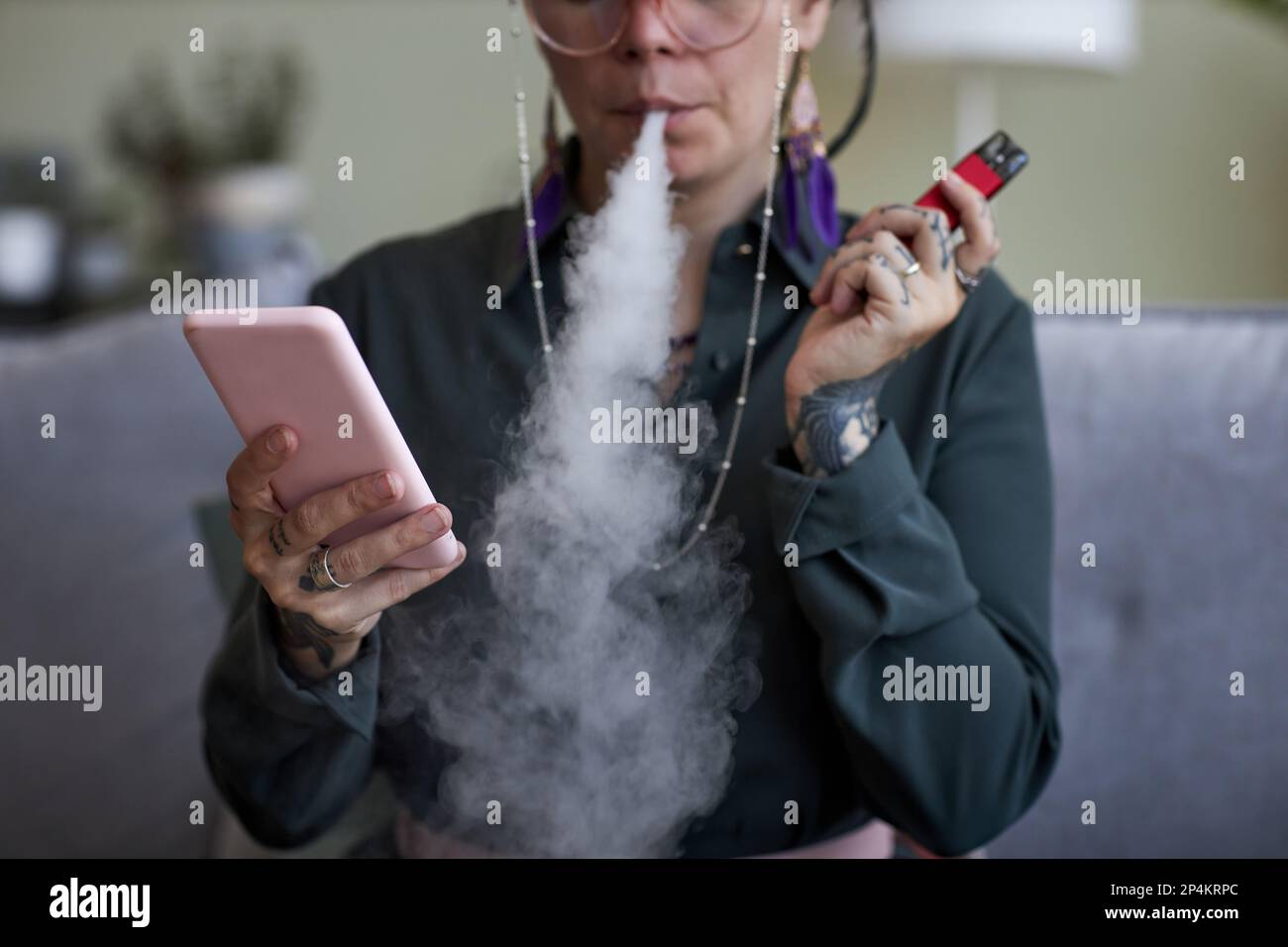 Close-up of young woman with e-cigarette sitting on couch in front of  camera in home environment and texting in smartphone Stock Photo - Alamy
