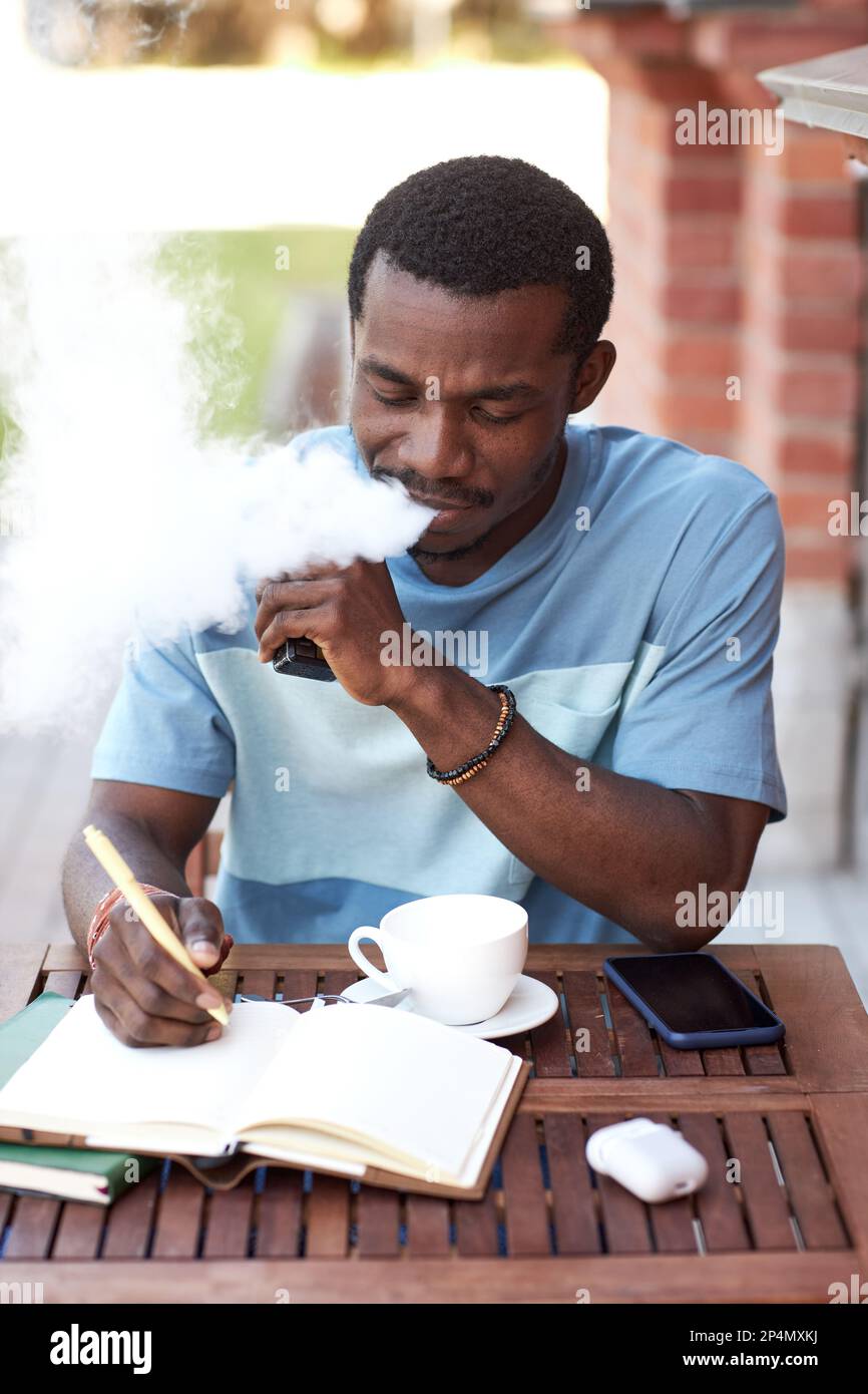 Young African American businessman smoking vape pen sitting by table in  outdoor cafe and making notes in notebook while organizing work Stock Photo 