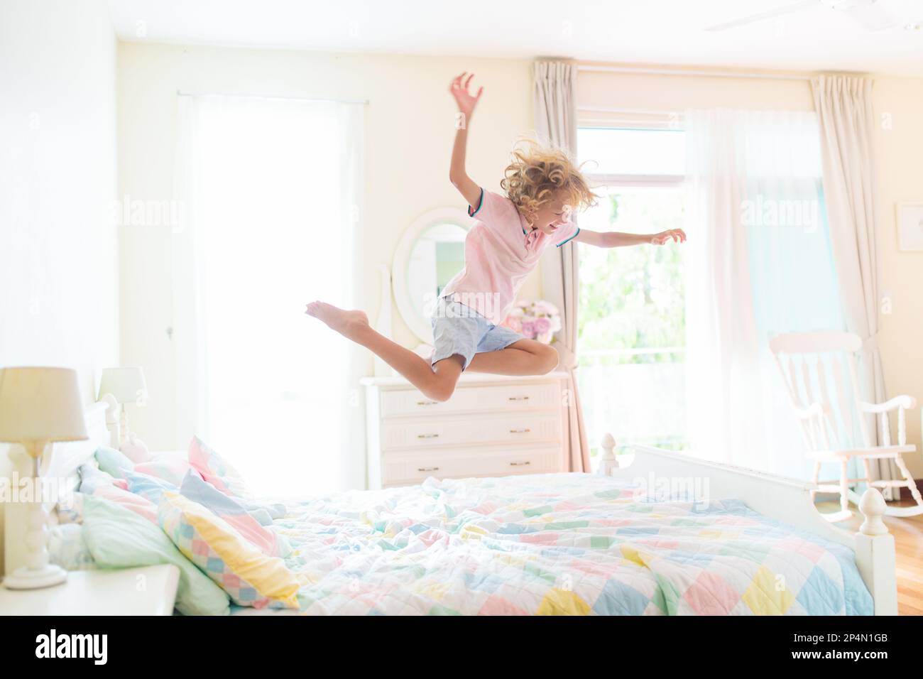 Child jumping on bed in white bedroom with big window. Kids play at