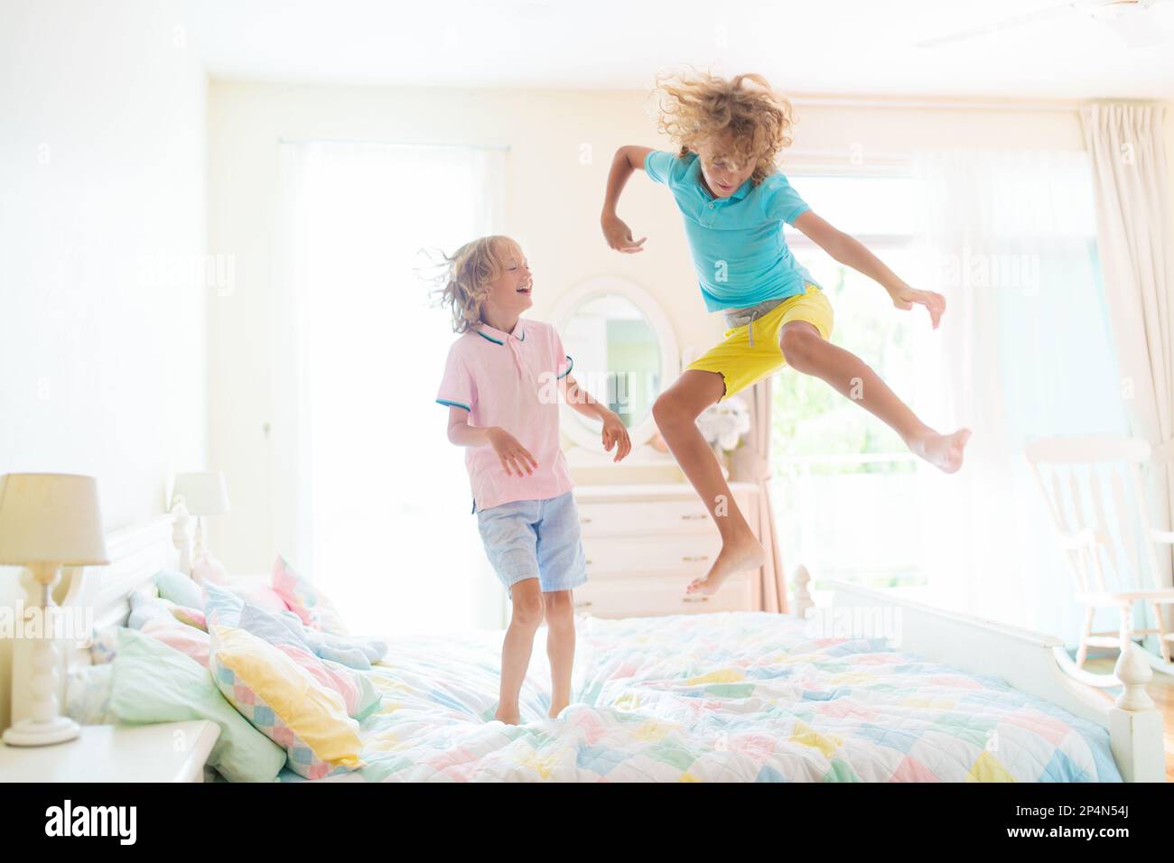 Child jumping on bed in white bedroom with big window. Kids play at