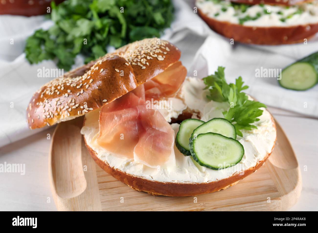 Delicious bagel with cream cheese, jamon, cucumber and parsley on wooden board, closeup Stock Photo