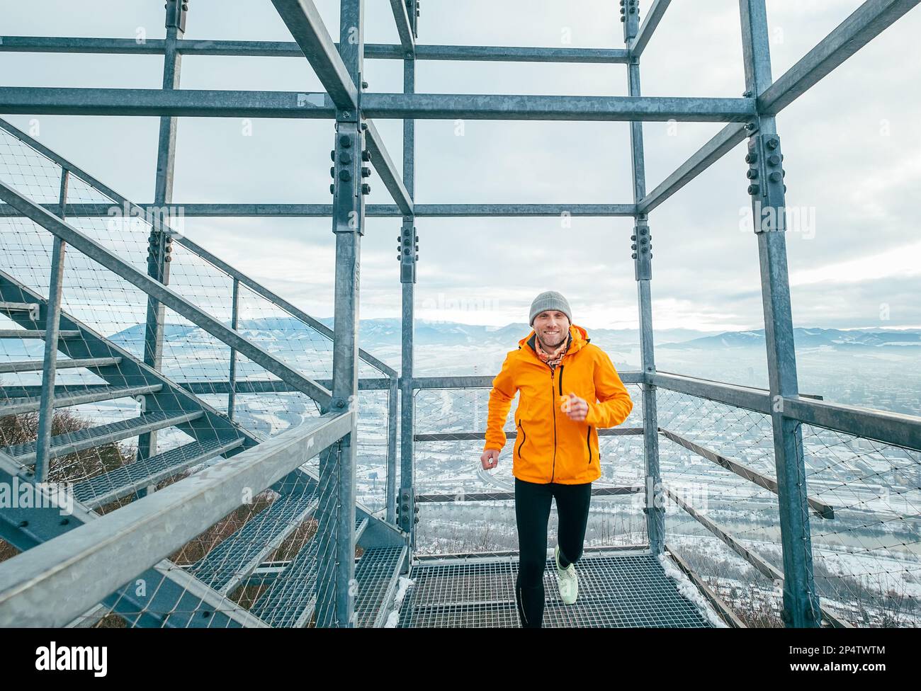 Smiling runner man dressed in bright orange softshell sporty clothes running down by huge steel industrial stairs in cold winter day. People healthy l Stock Photo