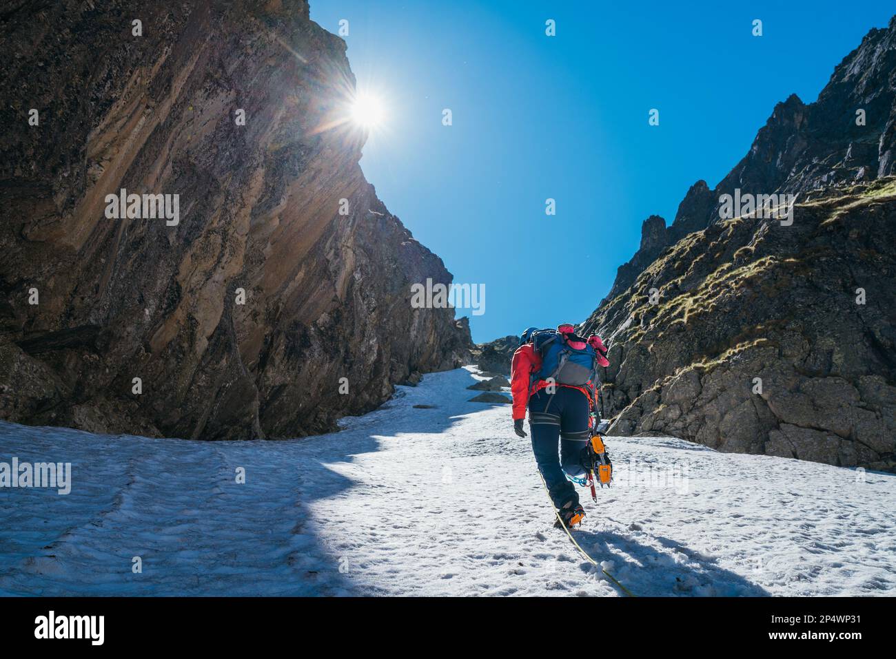 Team roping up woman dressed high altitude mountaineering clothes and harness climbing with backpack by snowy slope in the couloir with backlight sun Stock Photo