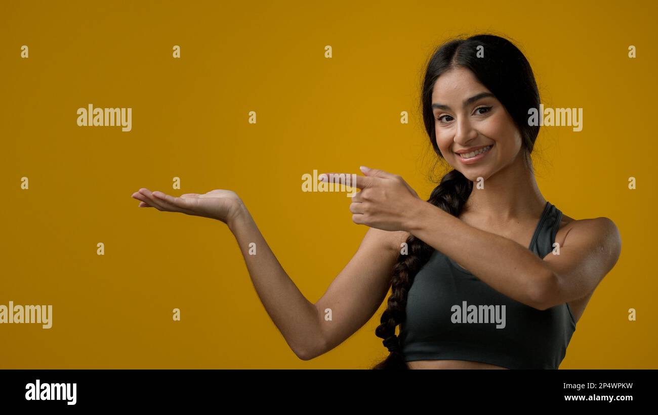 Portrait in studio Indian 20s woman posing on yellow background pointing to side at palm arm hand point aside recommend recommendation sign girl lady Stock Photo