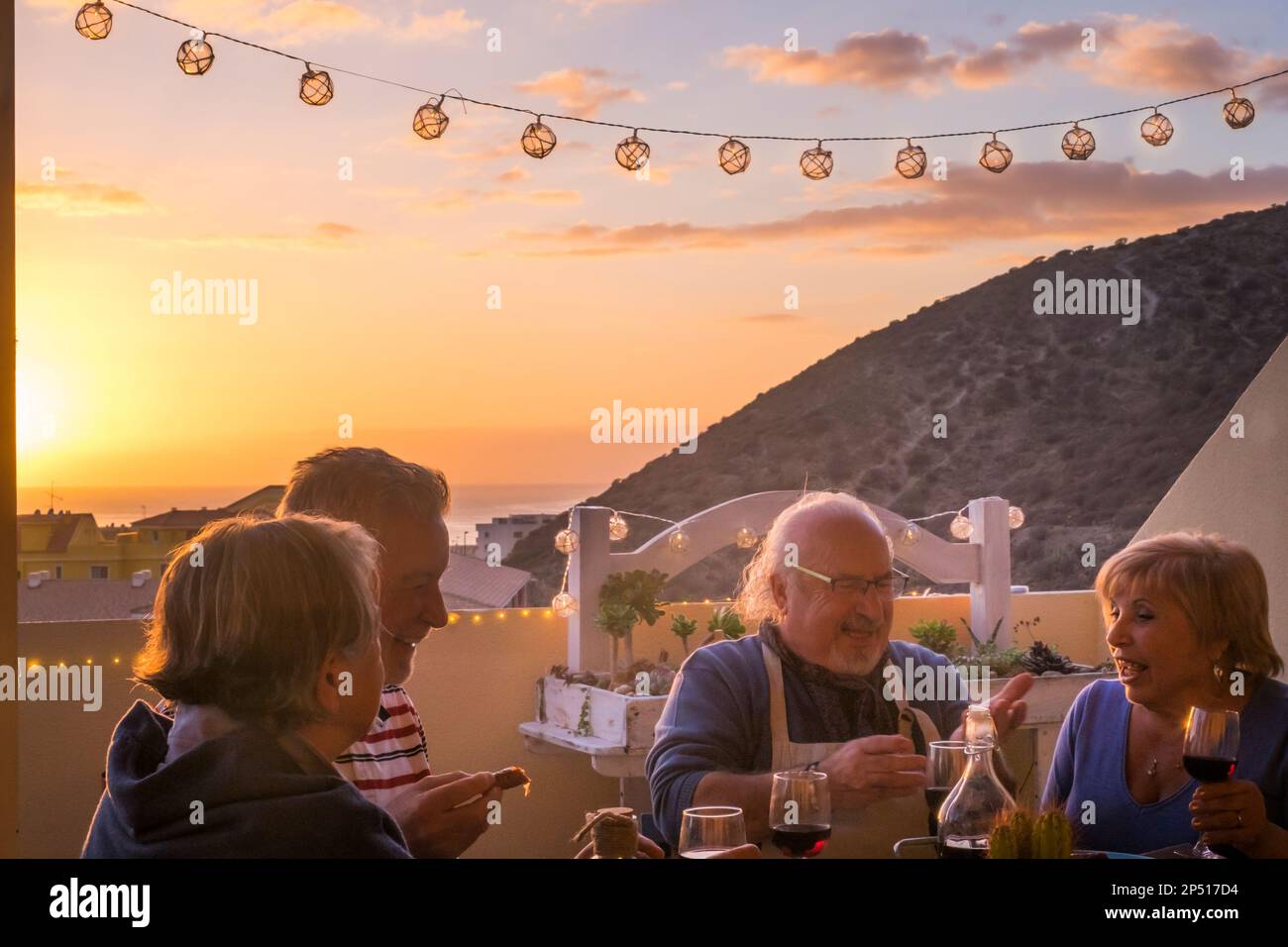 Happy group of senior people enjoy dinner time with sunset and town in background. Mature elderly lifestyle couples men and woman eating and drinking Stock Photo