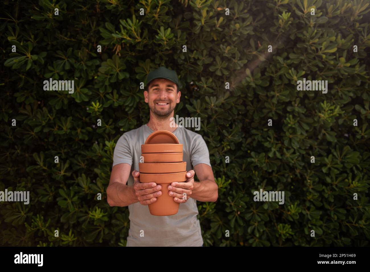 Close-up portrait man gardener in green cap holds terracotta clay pots in hands against the backdrop of an evergreen fence. Gardening, small business, Stock Photo