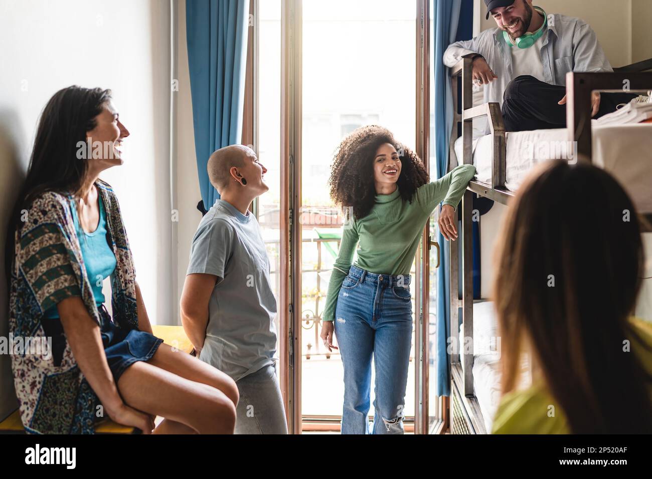 A group of diverse young adults relax in a hostel dorm room, laughing and chatting together. One individual has a shaved head, piercings, and androgyn Stock Photo