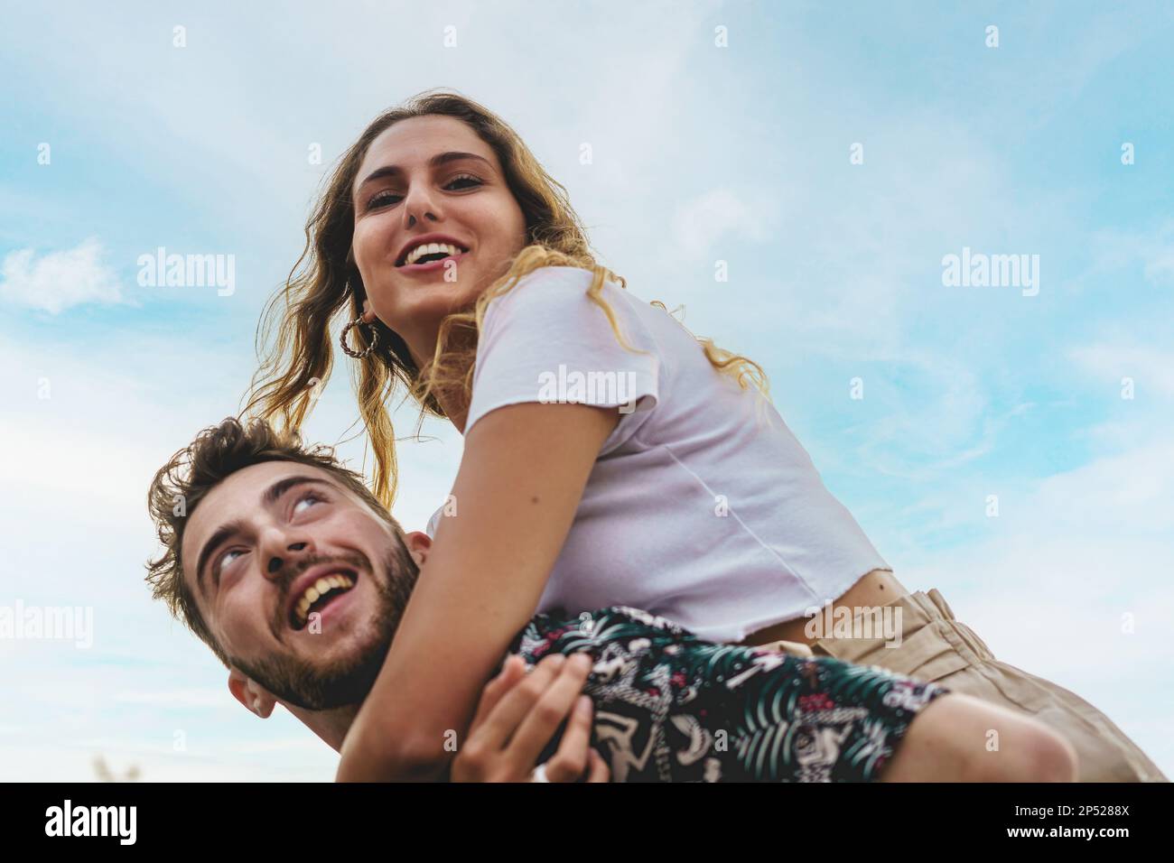 Two young adults, a Caucasian man and woman in their early twenties, are seen playfully embracing and laughing outdoors under a clear blue sky - young Stock Photo