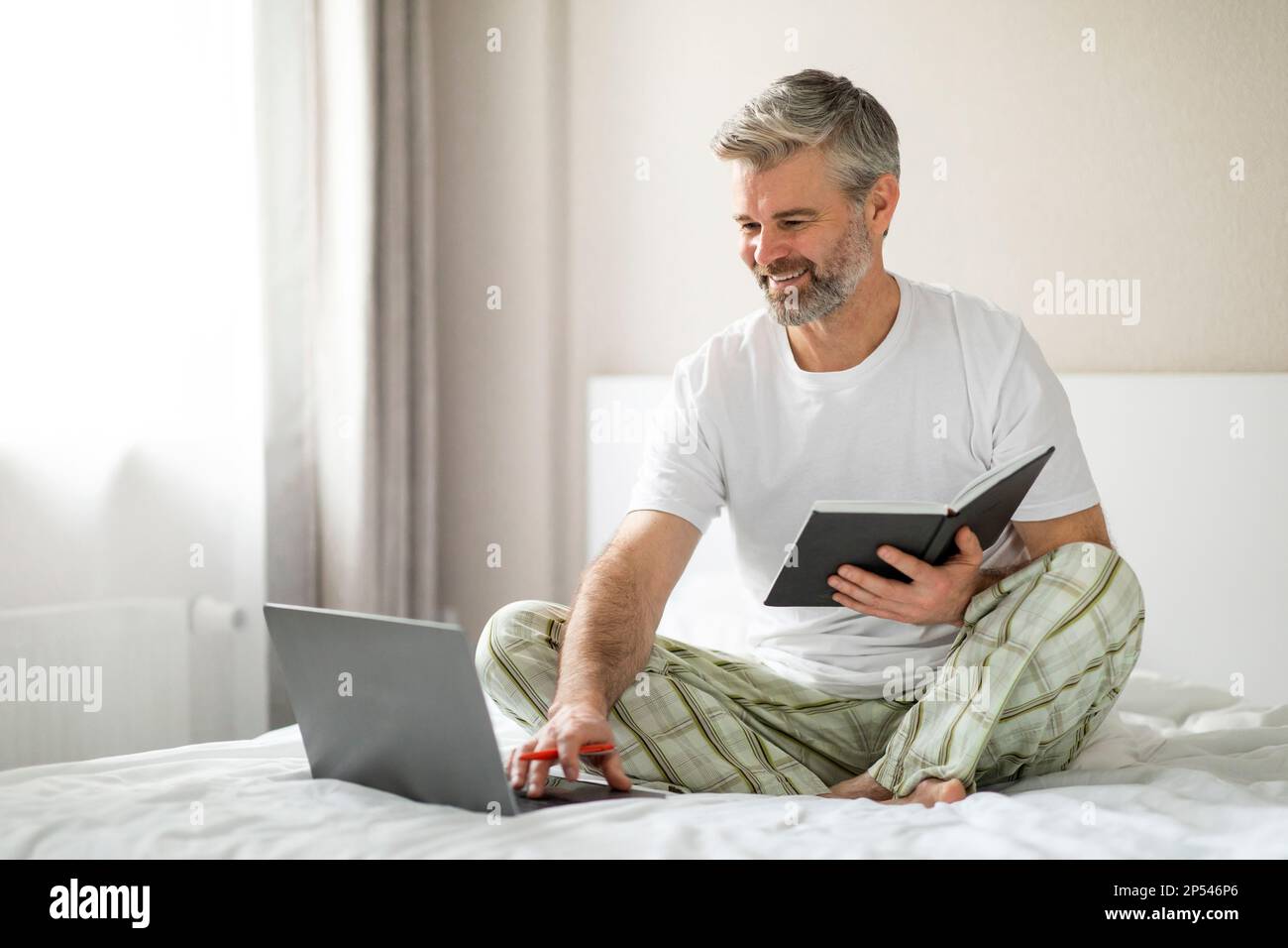 Cheerful mature man using laptop, taking notes in notepad Stock Photo