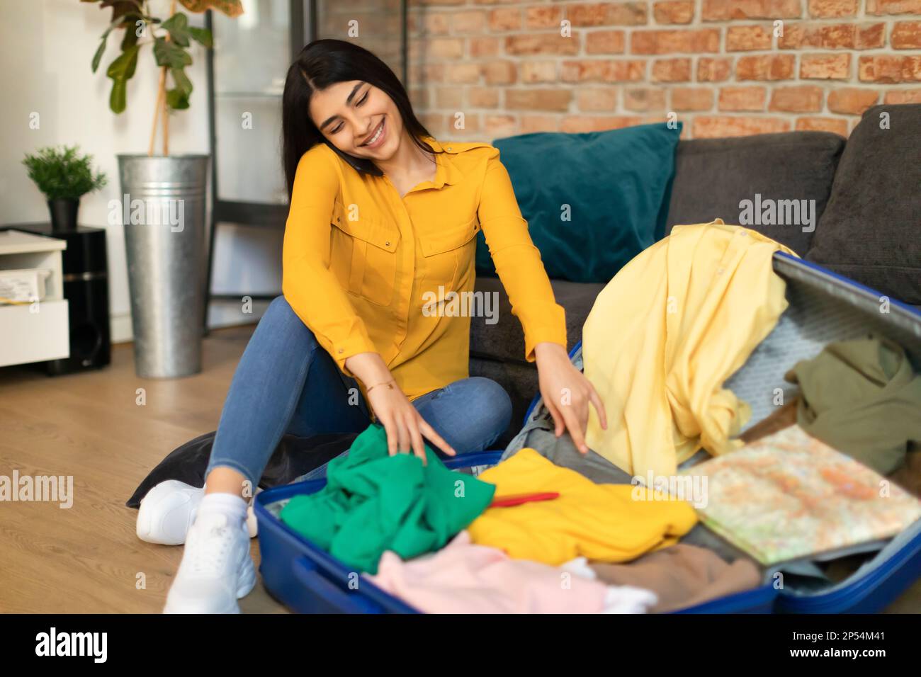 Happy mexican lady sitting near open suitcase with clothes, speaking on cellphone, making hotel reservation Stock Photo