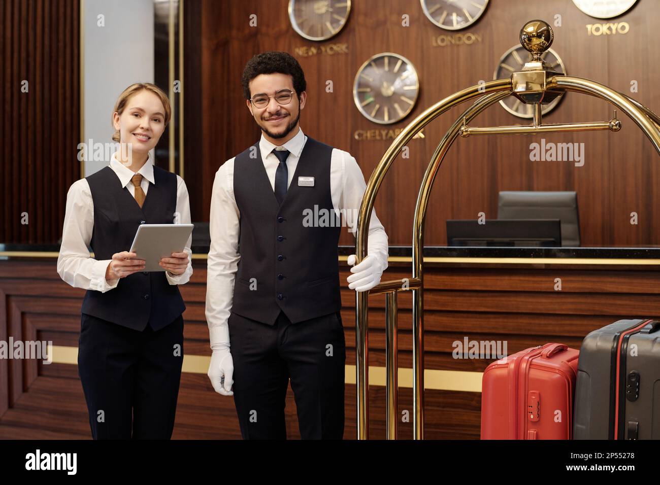 Happy young elegant receptionist and bellboy standing in lounge of luxurious hotel and looking at camera while waiting for new guests Stock Photo
