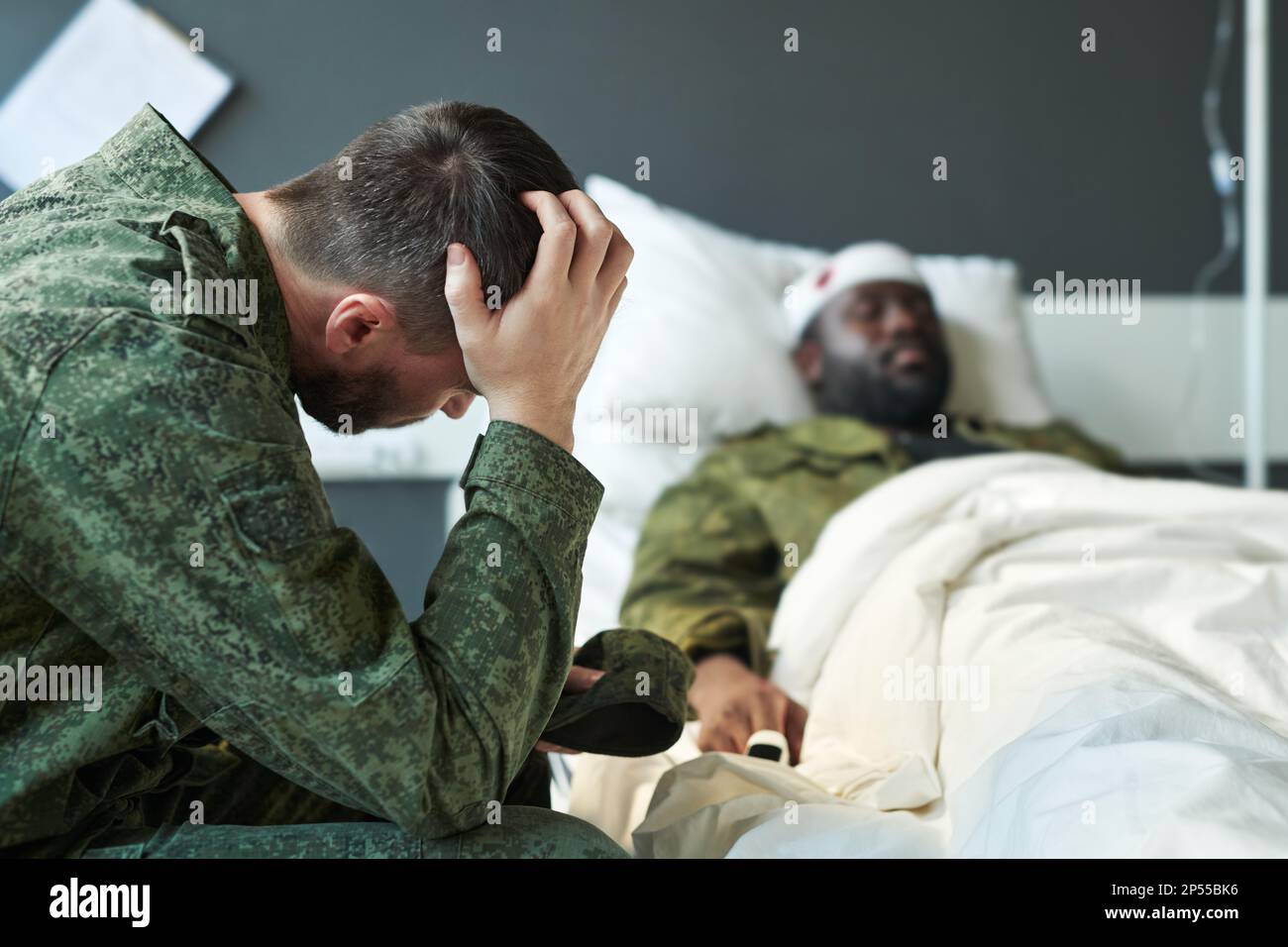 Young worried soldier in camouflage touching head while sitting by his friend in hospital bed after injury during military activities Stock Photo