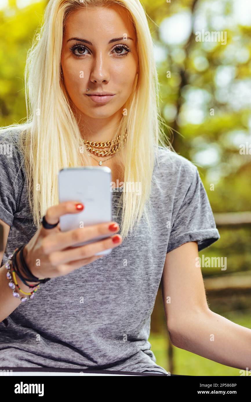 Beautiful blond girl in a park, has long hair and cell phone in hand. It could be summer or fall, but she is wearing a short-sleeved gray T-shirt. Stock Photo