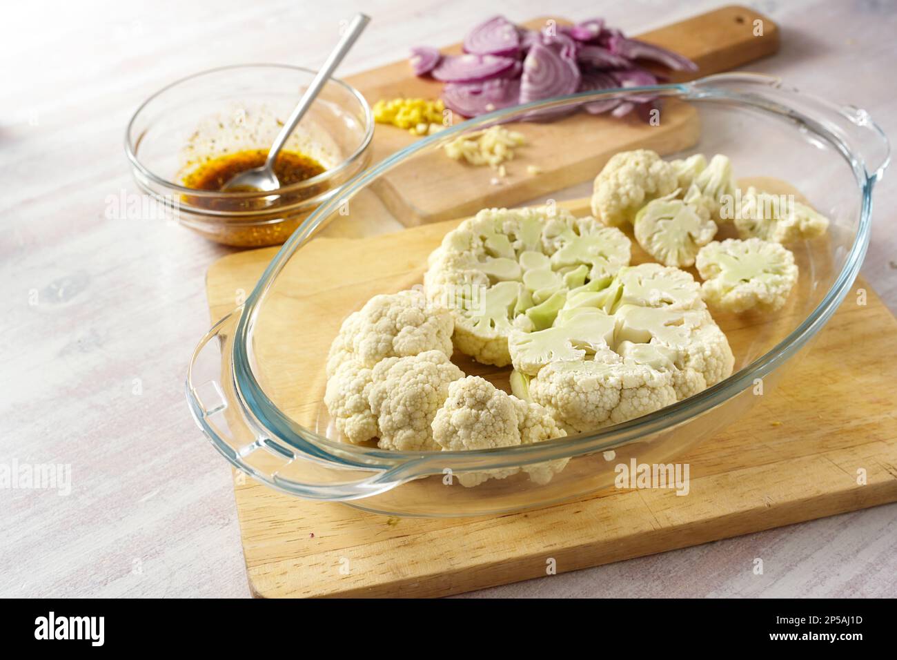 Raw cauliflower slices in a glass casserole, chopped red onion and a spice mix in olive oil, cooking preparation for baked vegetable steaks, copy spac Stock Photo