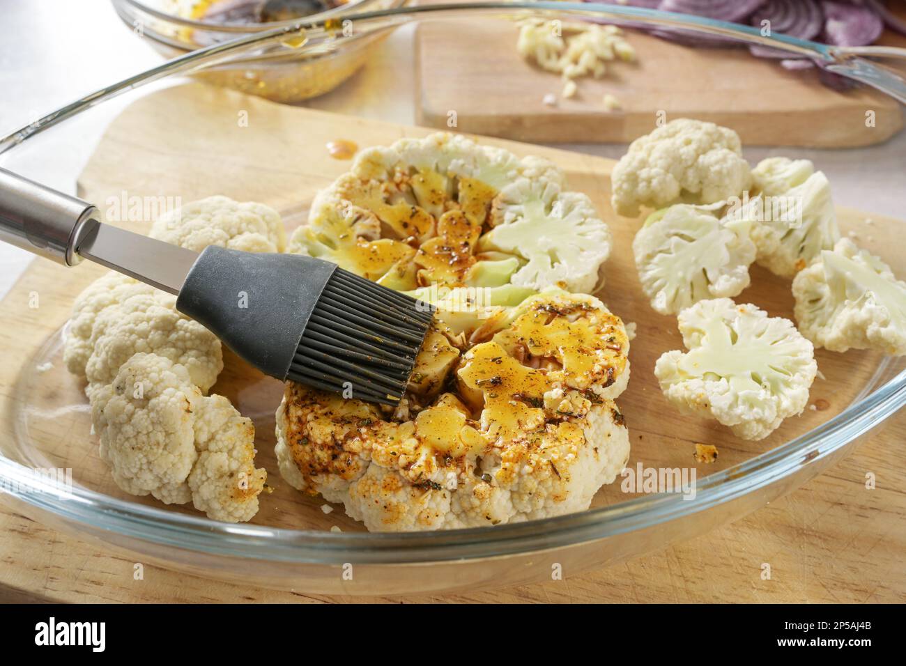 Raw cauliflower slices in a glass casserole are coated with a mixture of spices, herbs and olive oil by a silicone brush, cooking preparation for bake Stock Photo