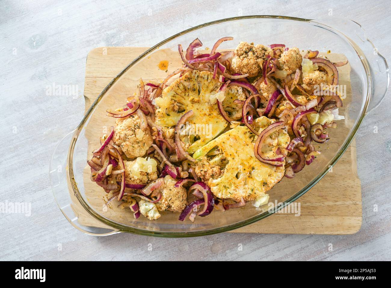 Baked cauliflower steaks with spicy oil and red onions in a glass casserole on a wooden kitchen board on a light table, vegetarian vegetable meal, top Stock Photo