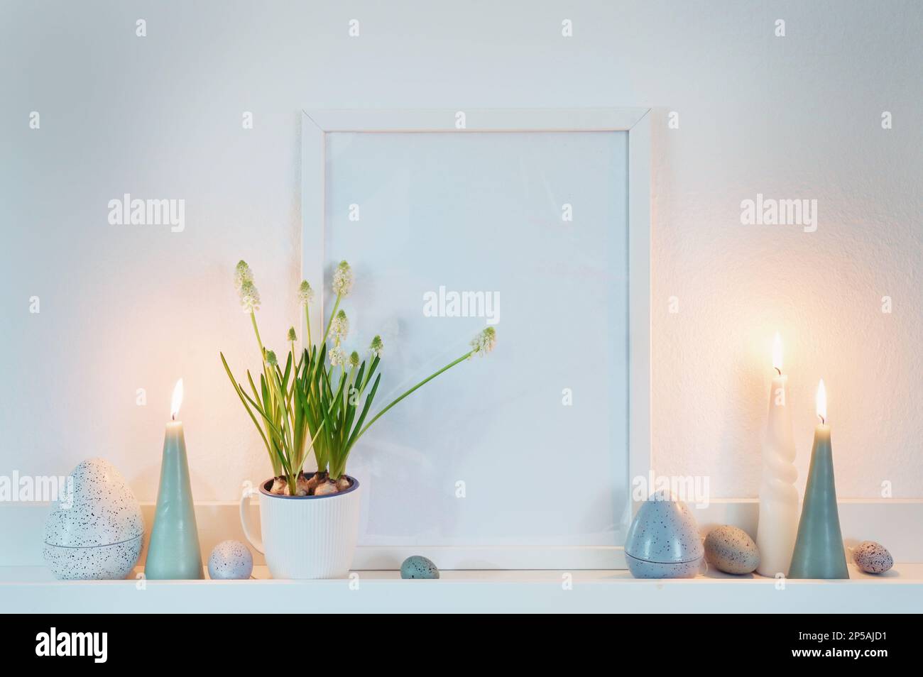 Decoration with an empty picture frame, candles and easter eggs in dusty blue green colors and a potted grape hyacinth plant on a white shelf on the w Stock Photo