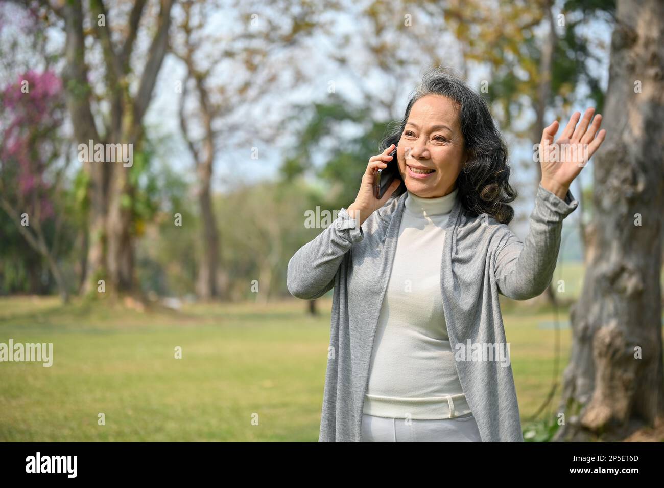 Happy aged Asian woman in casual clothes waving her hand and making a hello gesture, bumped into her neighbor in the green park while talking on the p Stock Photo