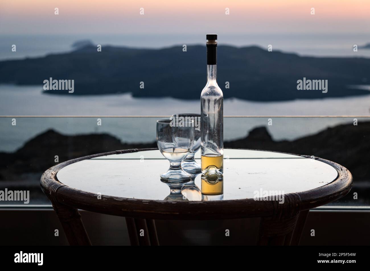 Dusk view from Fira town,  Santorini of glasses with bottle of raki or tsikoudia, a traditional Cretan spirit, a 100% natural product. Stock Photo