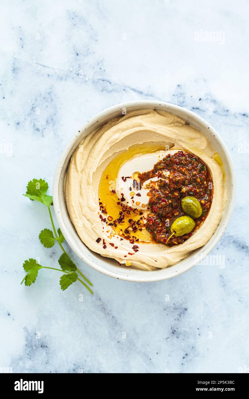 Homemade spicy hummus with tahini and olive oil in a bowl, white marble