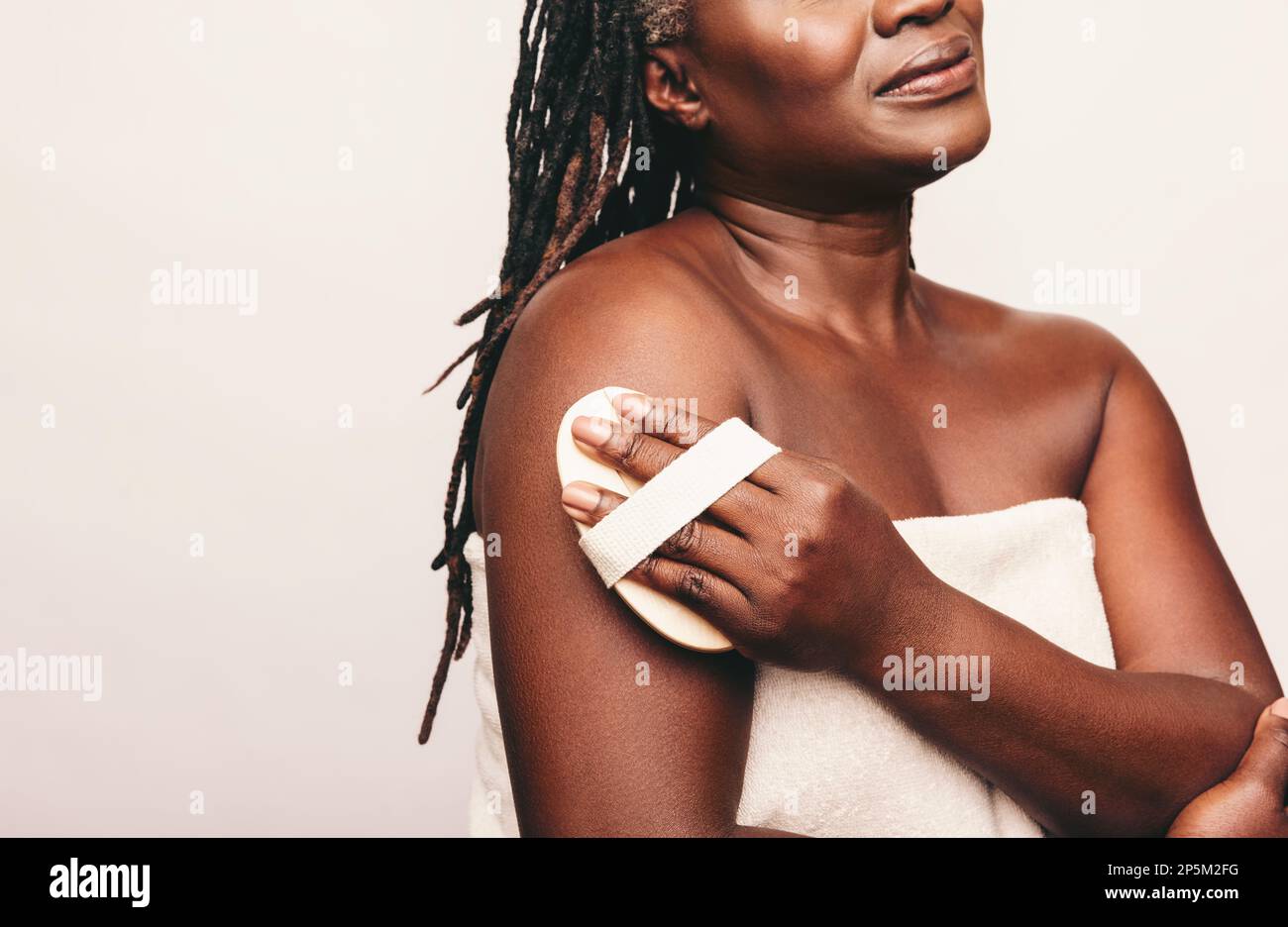 Mature woman exfoliating her skin with a brush against a studio background. Woman with dreadlocks dry brushing her arm while wrapped in a bath towel. Stock Photo