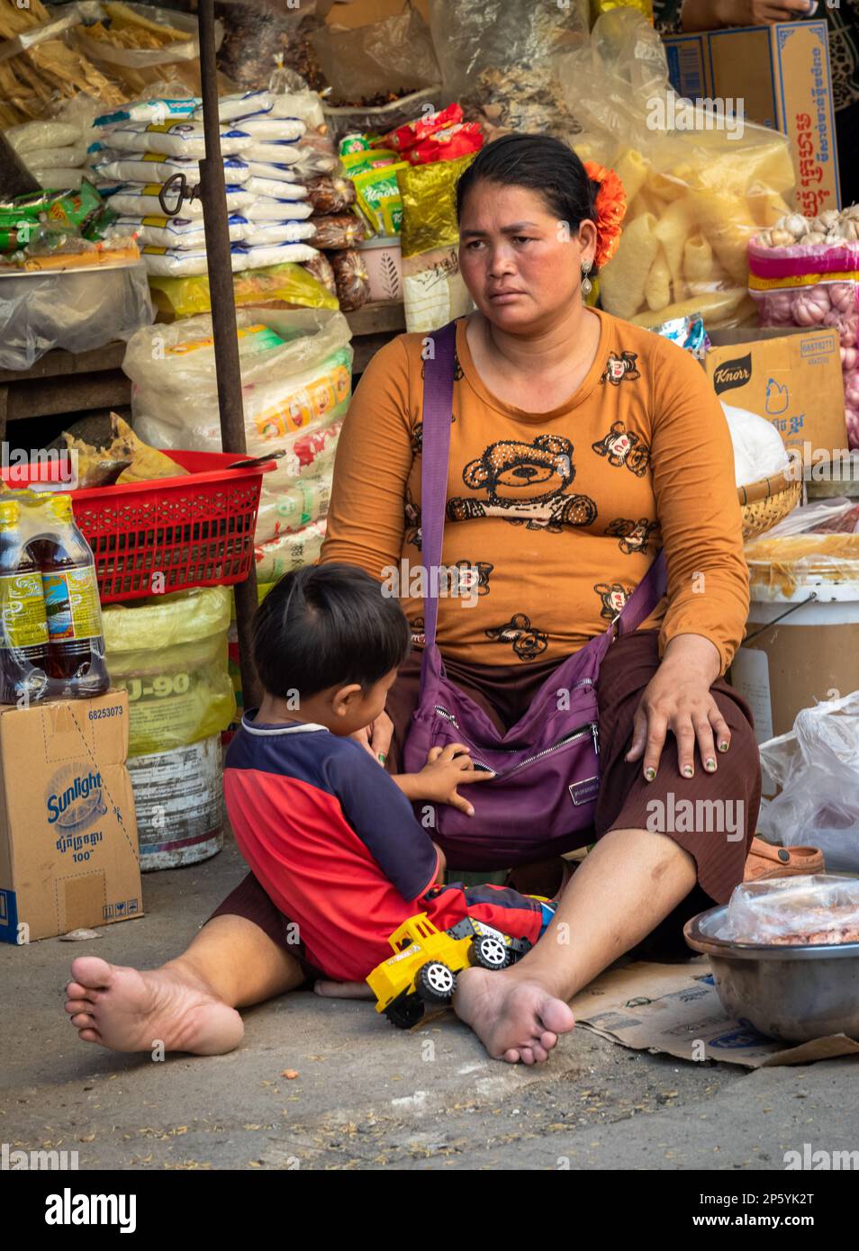 A female grocery stall holder in a street market in Phnom Penh, Cambodia, sits with her son by her feet and looks sad. Stock Photo