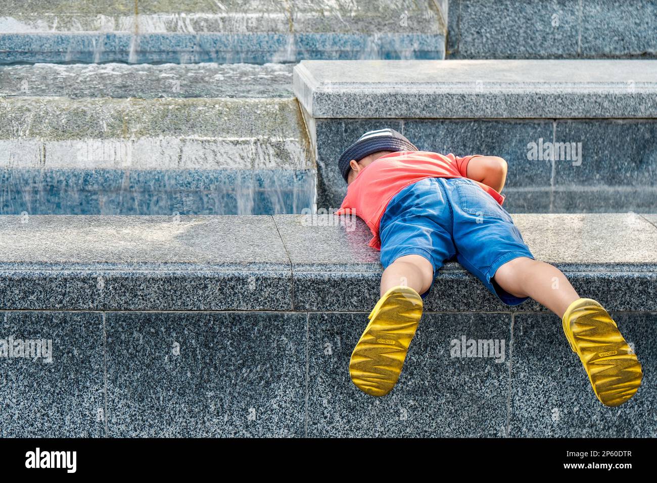 Funny little boy with yellow shoes tries to touch water in fountain lying on parapet backside view. Curious kid plays on city street Stock Photo