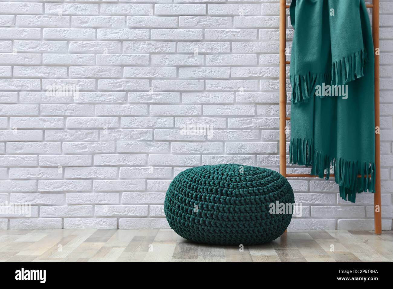 Stylish knitted pouf and ladder with plaid near white brick wall ...