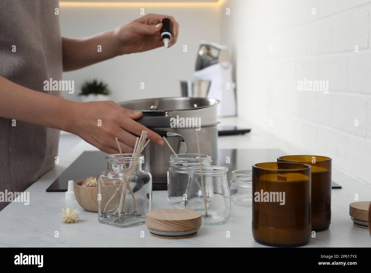 Woman adding essential oil into pot with melted wax on stove in kitchen
