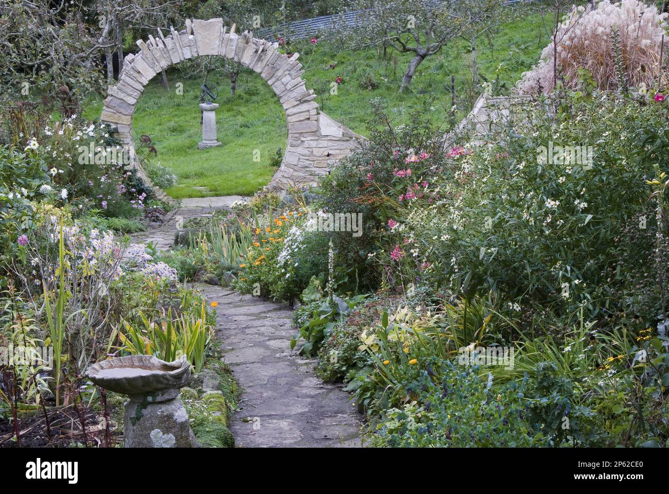 old stone paving path in country garden with flower borders leading to stone moon gate with garden view beyond Stock Photo