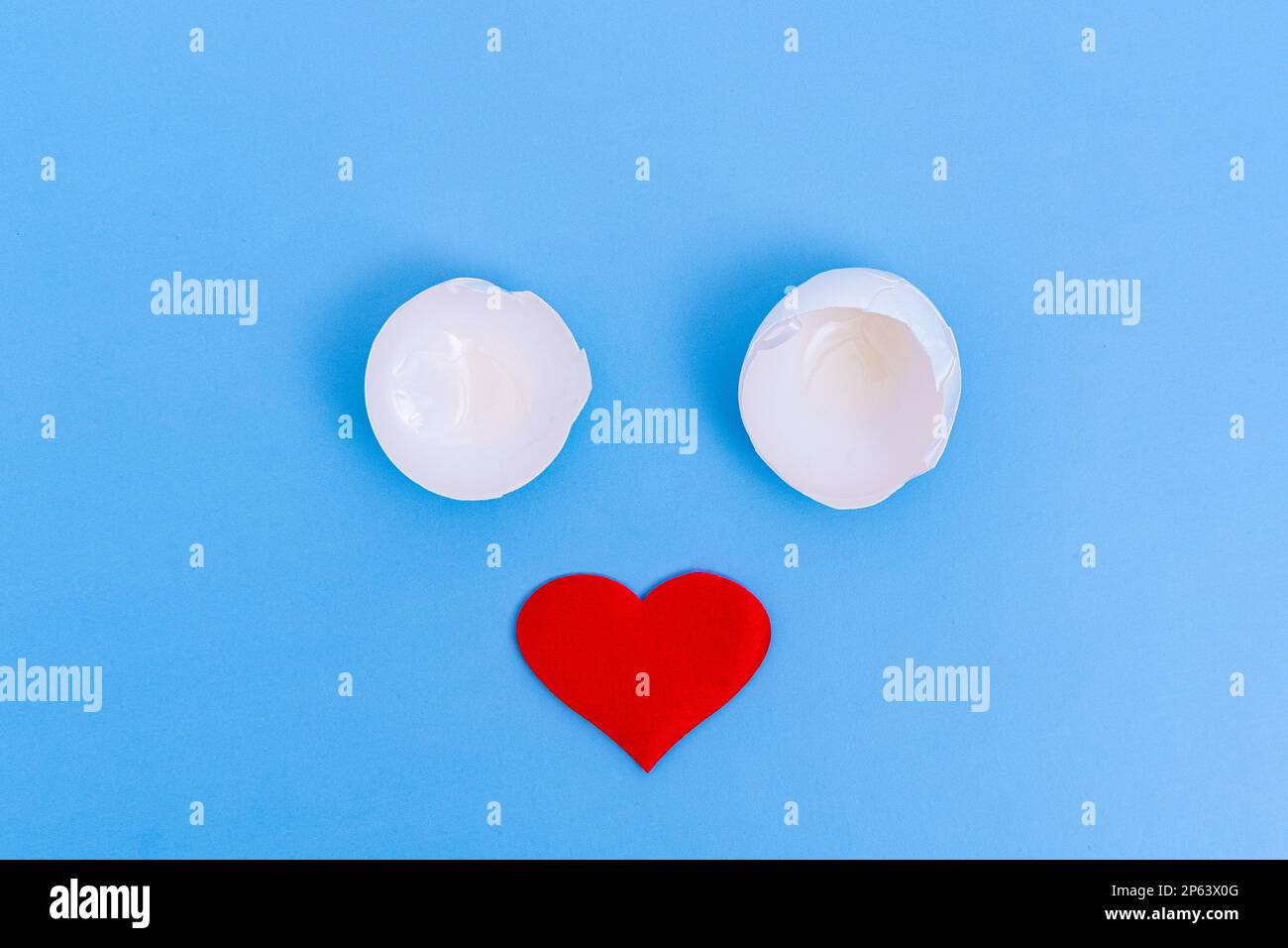 One is a white broken eggshell and a red heart on a blue background. A creative face made of two eggshells as eyes and a nose heart. Stock Photo