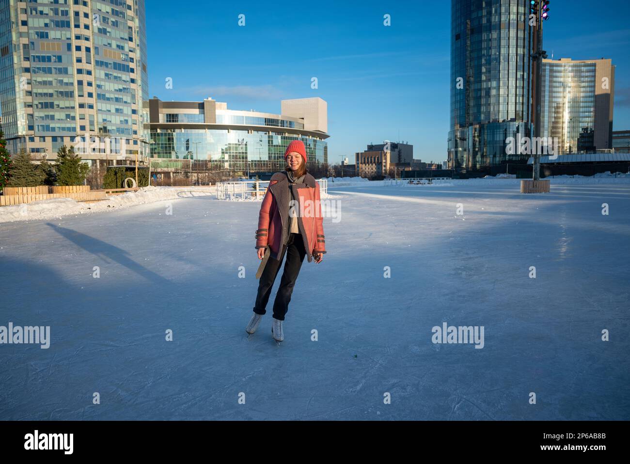 a young beautiful girl on skates in a sheepskin coat on a skating rink against the backdrop of the metropolis and the rays of the sun. Stock Photo