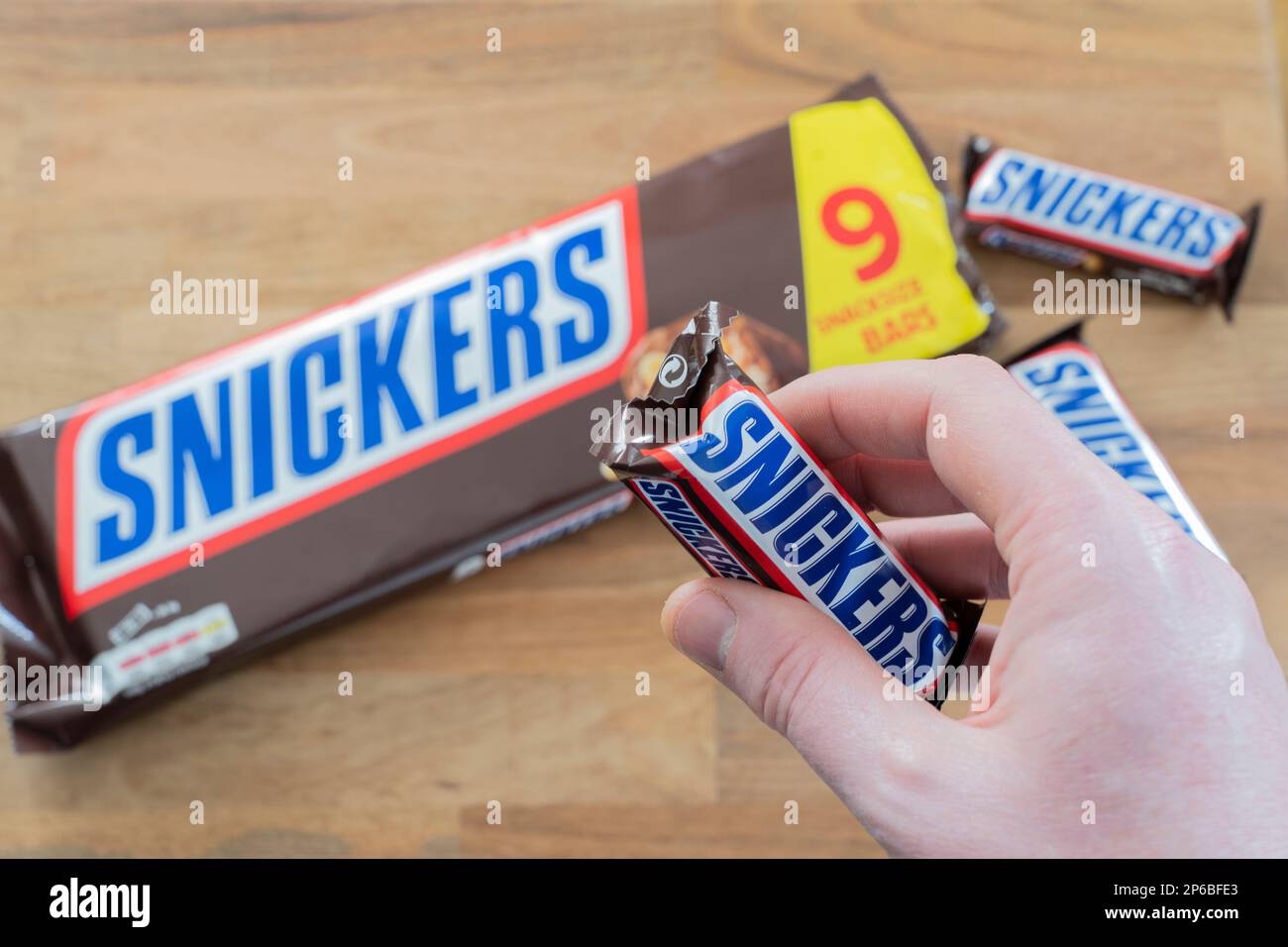 Hand holding a snickers bar with a snickers packet in the background. Snickers is owned by Mars, Incorporated. Theme: snacking, snack food, unhealthy Stock Photo