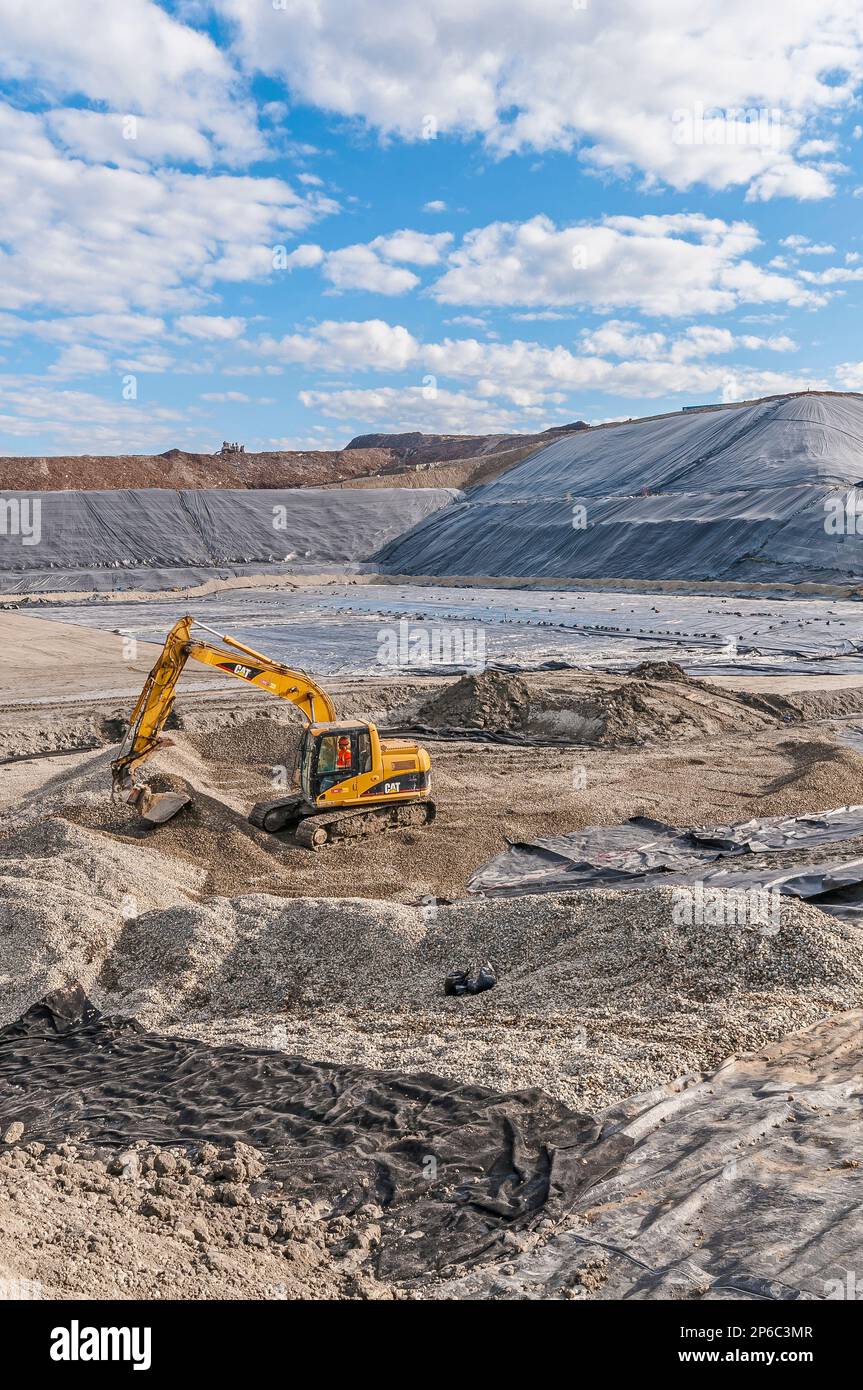 A worker in a power shovel digging in areas of excavation and plastic geomembrane coverings at an active landfill. Stock Photo