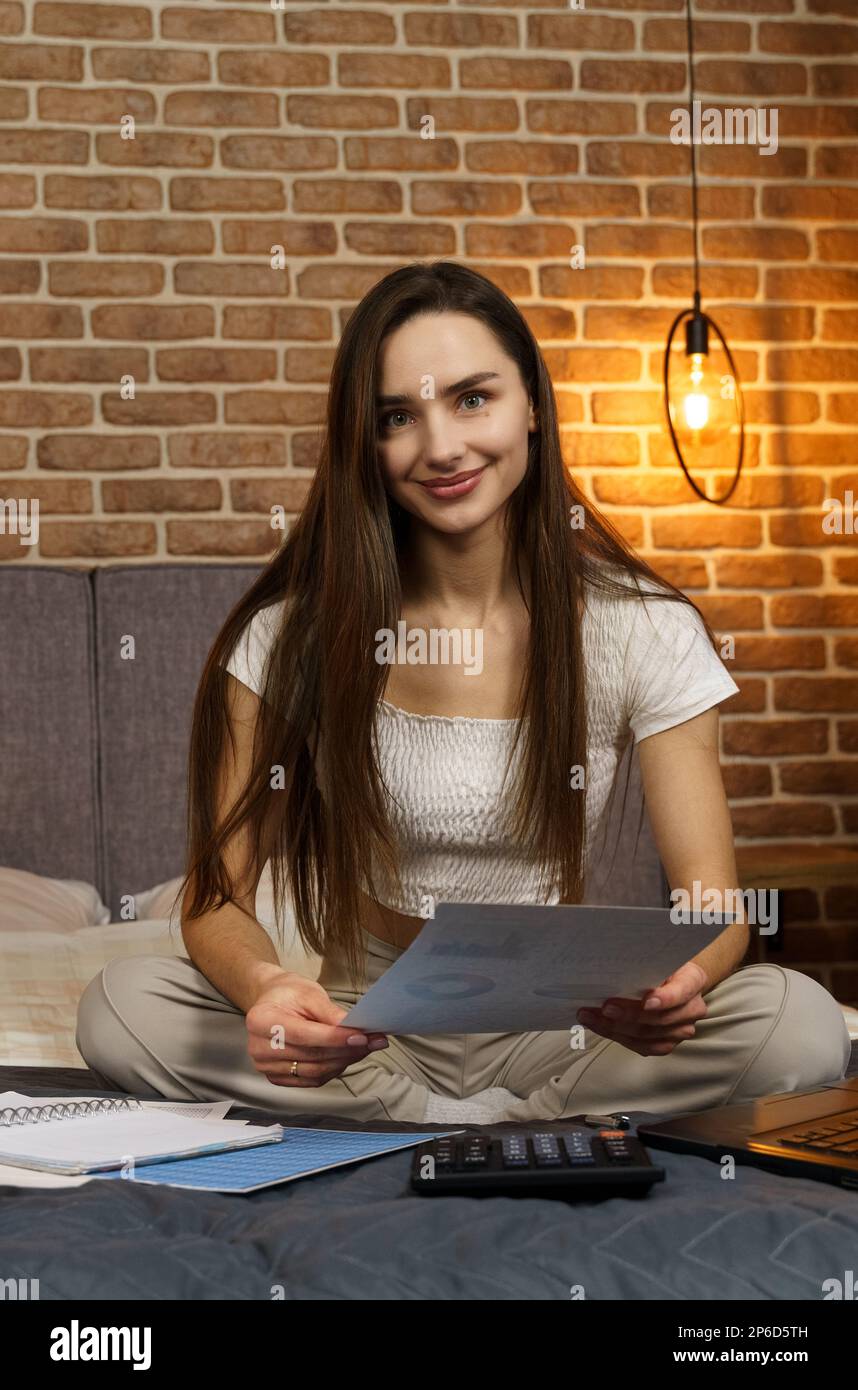 A young woman sits on a bed, looks at reporting documents, counts on a