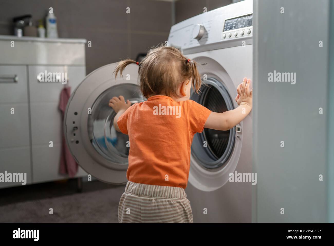One girl small caucasian toddler child daughter standing at the washing machine in the toilet opening or closing the door examine and learn early deve Stock Photo