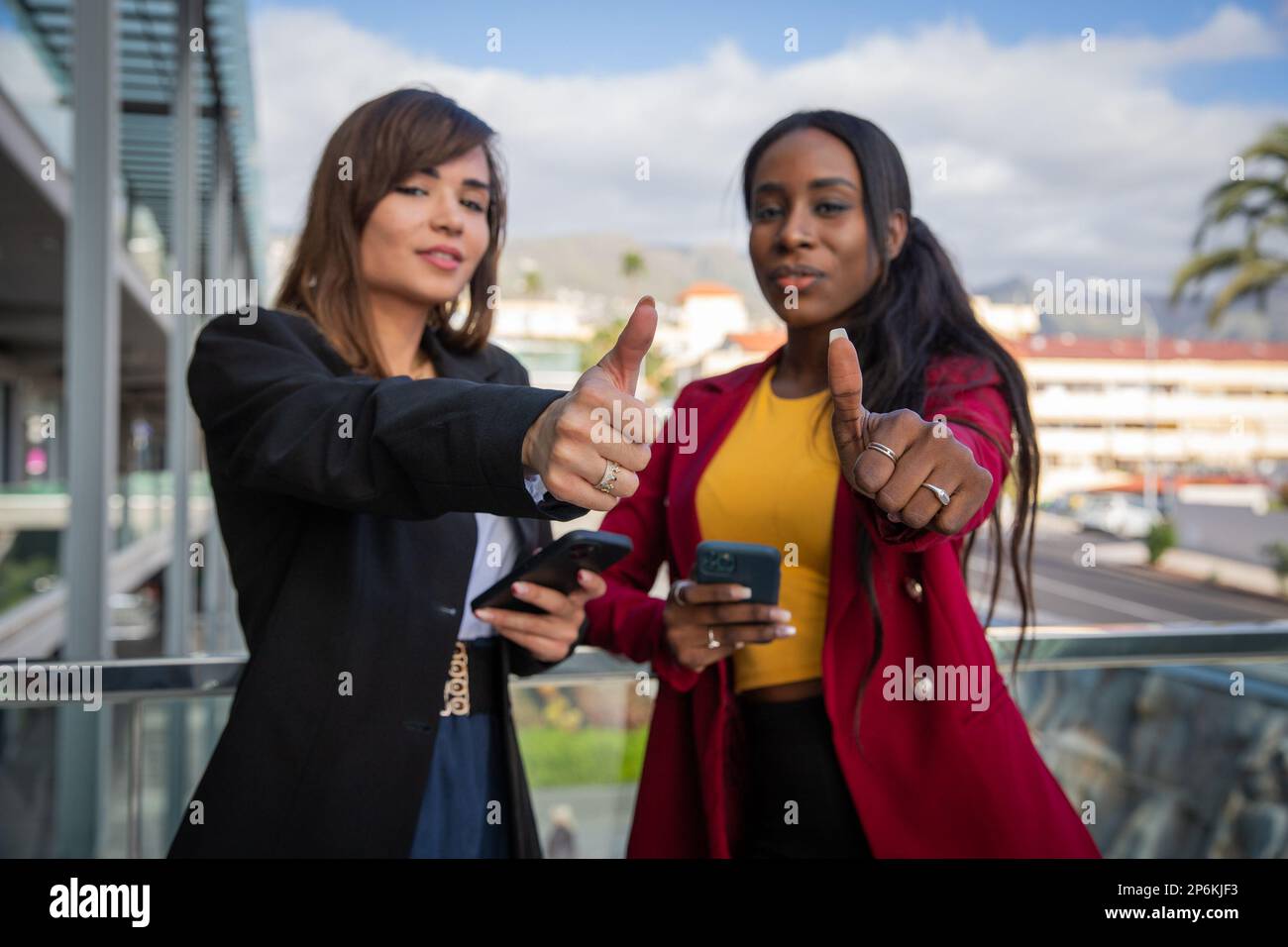 Two female colleagues show thumbs up while working using their smartphones Stock Photo