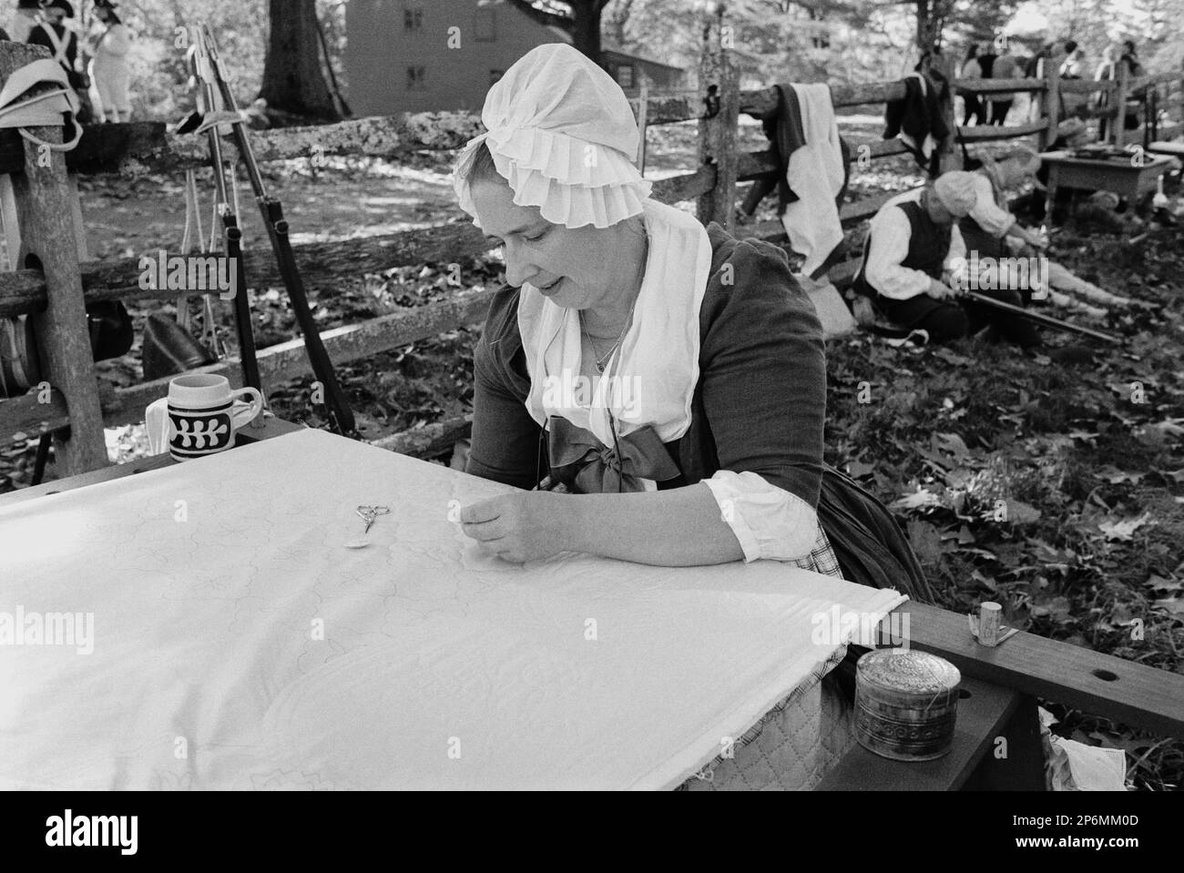 An actress in period clothing works on sewing a complex quilt outdoors while awaiting a battle  during a Revolutionary War reenactment at the Rebecca Stock Photo